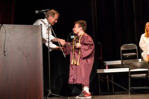 Young man in a maroon graduation gown standing on a stage holding a award next to a podium and shaking hands with an older man