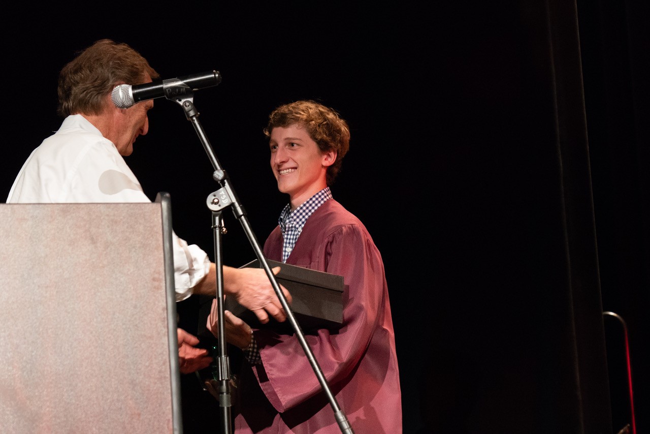 High school aged boy being handed an award by an older man and smiling on a stage. He is wearing a maroon graduation gown.